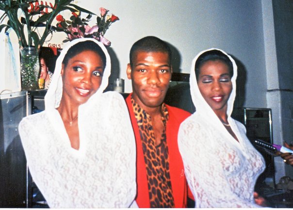 Three Black people, Roshumba, Douglas Says, and Danielle Revlon, two wearing white lace with hoods and Douglas wearing a red jacket and a leopard print dress shirt.