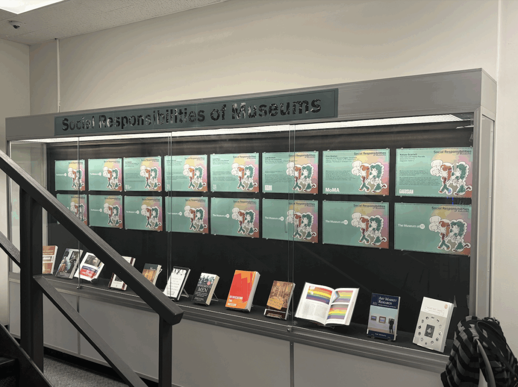 A photograph of the 4th-floor display case in the FIT library, showing the exhibit Social Responsibilities of Museums. The case contains printed graphics and a selection of books.