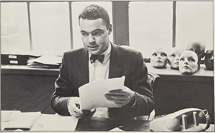 Black and white photograph of Colin Birch in his office at Bloomingdales, with mannequin heads behind him. He has short dark hair, is wearing a jacket and bow tie, and is holding some papers.