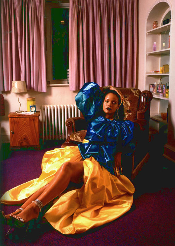 Photo of Rihanna lounging in pink room with blue blouse and yellow skirt.